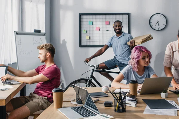 Selective focus of african american businessman on bike holding pizza boxes near multicultural colleagues working in office — Stock Photo