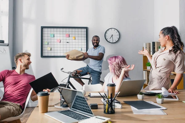 Selective focus of business people looking at african american colleague on bike with takeaway pizza in office — Stock Photo