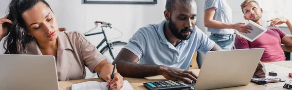 Orientação panorâmica de empresária escrevendo em notebook perto de colega afro-americano usando laptop no escritório — Fotografia de Stock