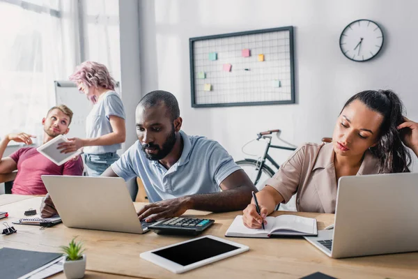 Enfoque selectivo de la mujer de negocios escribiendo en el cuaderno, mientras que el hombre de negocios afroamericano utilizando el ordenador portátil en la mesa en la oficina - foto de stock