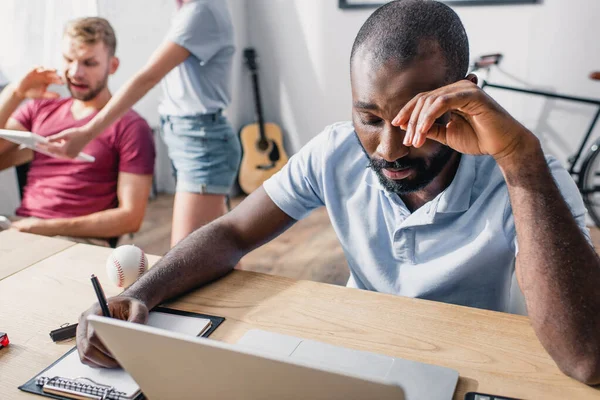 Focus selettivo di stanco uomo d'affari africano americano che scrive sugli appunti vicino al computer portatile sul tavolo in ufficio — Foto stock