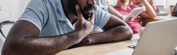 Cultivo panorámico de hombre de negocios afroamericano sentado cerca de la computadora portátil en la mesa de oficina - foto de stock