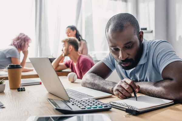 Selektiver Fokus eines afrikanisch-amerikanischen Geschäftsmannes, der mit Klemmbrett, Taschenrechner und Laptop auf dem Bürotisch arbeitet — Stockfoto