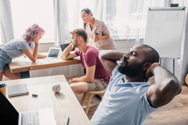 Selektiver Fokus eines afrikanisch-amerikanischen Geschäftsmannes, dessen Hände in Kopfnähe am Tisch sitzen, während multikulturelle Kollegen im Büro arbeiten — Stockfoto