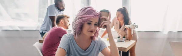 Panoramische Ernte verärgerter Geschäftsfrau mit Pappbecher, während multikulturelle Kollegen im Hintergrund im Büro arbeiten — Stockfoto