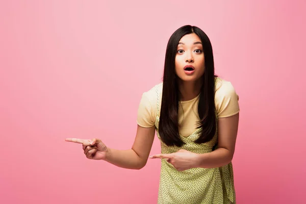 Conmocionada chica asiática señalando con los dedos y mirando a la cámara aislada en rosa - foto de stock