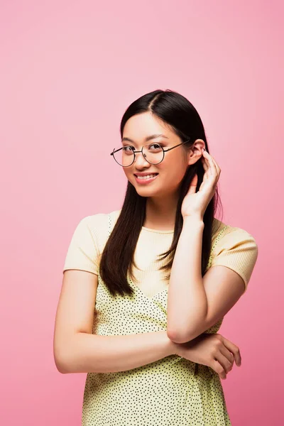 Cheerful asian girl in glasses touching hair isolated on pink — Stock Photo