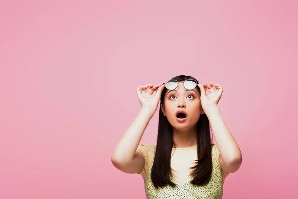 Shocked asian girl looking up and touching glasses isolated on pink — Stock Photo