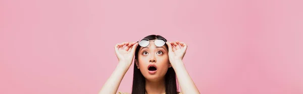 Plan panoramique de choqué asiatique fille regardant vers le haut et touchant lunettes isolé sur rose — Photo de stock