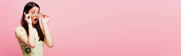 Panorâmica tiro de chocado asiático menina olhando afastado e tocando óculos isolado no rosa — Fotografia de Stock