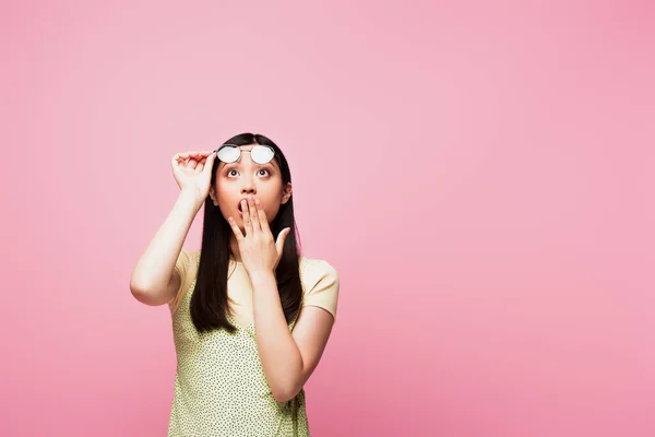 Conmocionada chica asiática mirando hacia arriba, cubriendo la boca y tocando gafas aisladas en rosa - foto de stock
