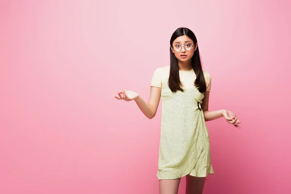 Verwirrtes asiatisches Mädchen mit Brille blickt in die Kamera und gestikuliert auf rosa — Stockfoto
