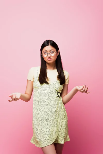 Mécontent asiatique fille dans lunettes montrant hausser les épaules geste sur rose — Photo de stock