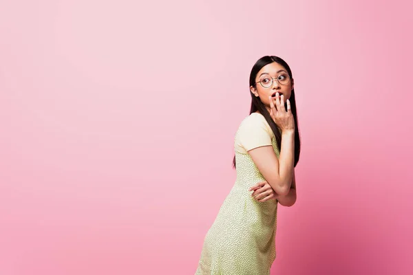 Jeune femme asiatique dans des lunettes couvrant la bouche et regardant loin sur rose — Photo de stock