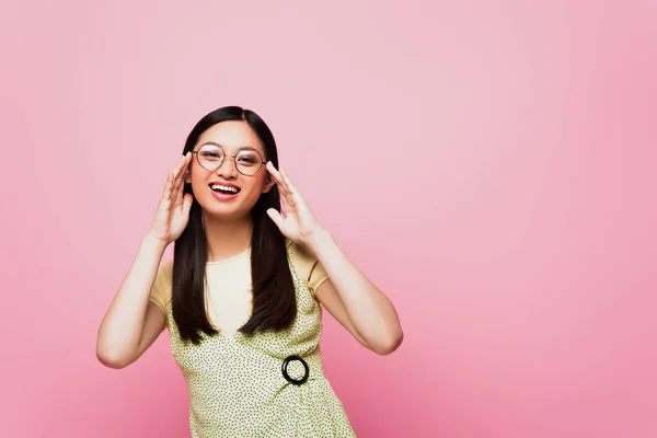 Fröhliche und junge asiatische Frau berührt Brille und lächelt auf rosa — Stockfoto