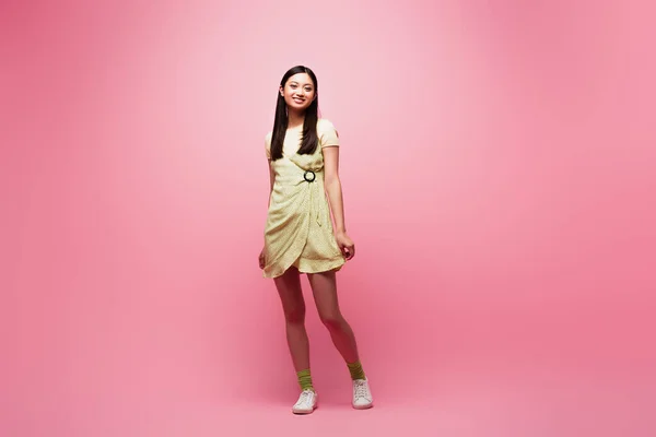 Cheerful asian young woman in dress standing on pink — Stock Photo