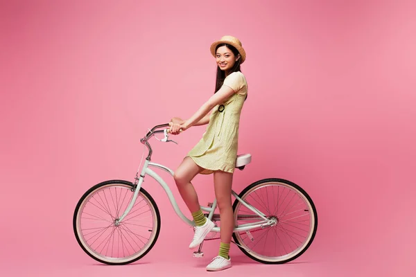 Весела азіатська молода жінка в солом'яному капелюсі біля велосипеда на рожевому — стокове фото
