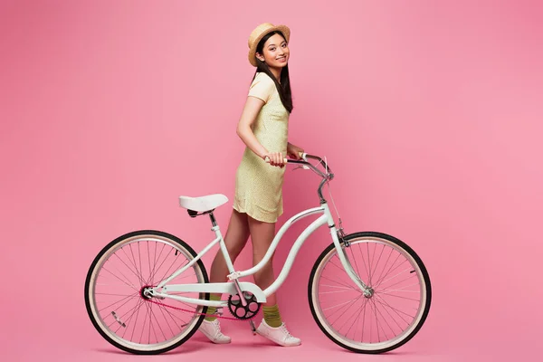 Fröhliche asiatische junge Frau mit Strohhut geht mit Fahrrad auf rosa — Stockfoto