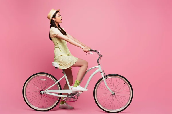 Lado vista de chocado ásia jovem mulher no palha chapéu equitação bicicleta no rosa — Fotografia de Stock