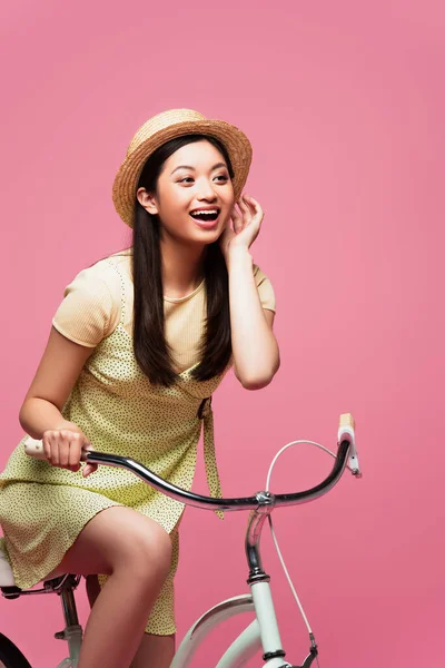 Emocionado asiático joven mujer en paja sombrero a caballo bicicleta en rosa - foto de stock