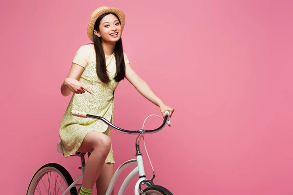 Smiling asian young woman in straw hat riding bicycle isolated on pink — Stock Photo