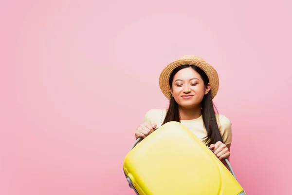 Animado ásia menina com fechado olhos no palha chapéu segurando bagagem isolado no rosa — Fotografia de Stock
