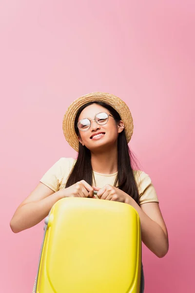 Позитивна азіатська дівчина в окулярах і солом'яному капелюсі тримає жовтий багаж на рожевому — стокове фото