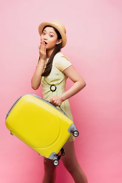 Shocked asian girl in straw hat holding yellow luggage and covering mouth on pink — Stock Photo
