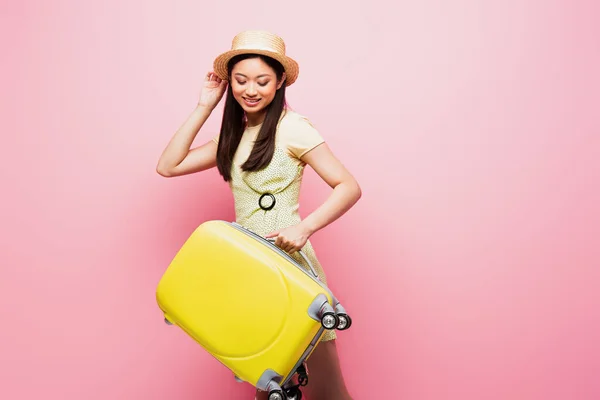 Positive asiatische Mädchen mit Strohhut Blick auf gelbes Gepäck auf rosa — Stockfoto