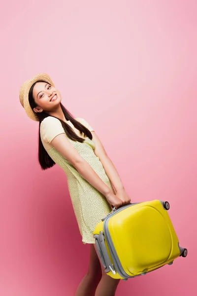 Позитивна азіатська дівчина в солом'яному капелюсі дивиться на камеру і тримає жовтий багаж на рожевому — стокове фото