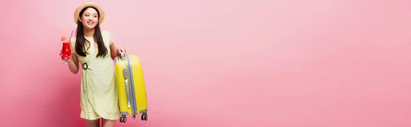 Panoramic concept of happy asian girl in straw hat holding cocktail and yellow baggage on pink — Stock Photo