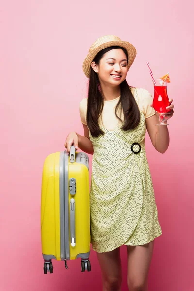 Feliz menina asiática em chapéu de palha segurando coquetel e bagagem amarela em rosa — Fotografia de Stock