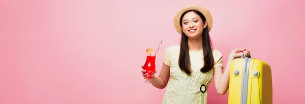 Site tête de heureux asiatique fille en paille chapeau tenant cocktail et jaune bagages sur rose — Photo de stock