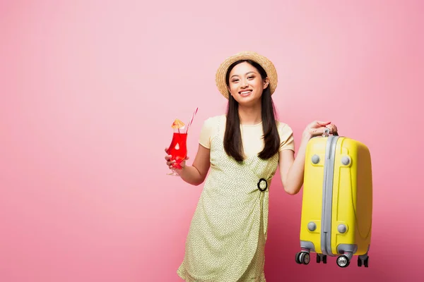 Sonriente asiático mujer en paja sombrero celebración cóctel y amarillo equipaje en rosa - foto de stock