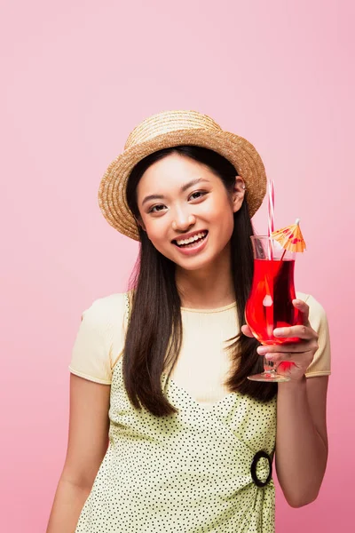 Sonriente asiático mujer en paja sombrero celebración vidrio con cóctel aislado en rosa - foto de stock