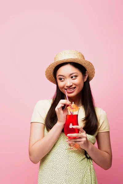 Sonriente asiático mujer en paja sombrero celebración vidrio con cóctel y mirando lejos aislado en rosa - foto de stock