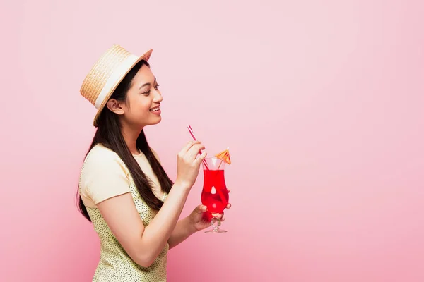 Усміхнена молода азіатка в солом'яному капелюсі тримає коктейль ізольовано на рожевому — стокове фото