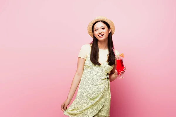 Cheerful young asian woman in straw hat and dress holding glass with cocktail on pink — Stock Photo