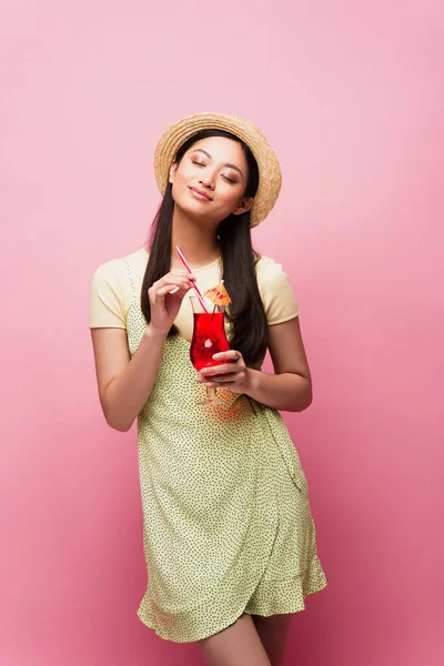 Sorrindo jovem mulher asiática com olhos fechados de pé e segurando vidro com coquetel em rosa — Fotografia de Stock