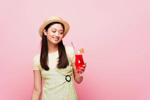 Sonriente joven asiático mujer en paja sombrero mirando vidrio con cóctel aislado en rosa - foto de stock