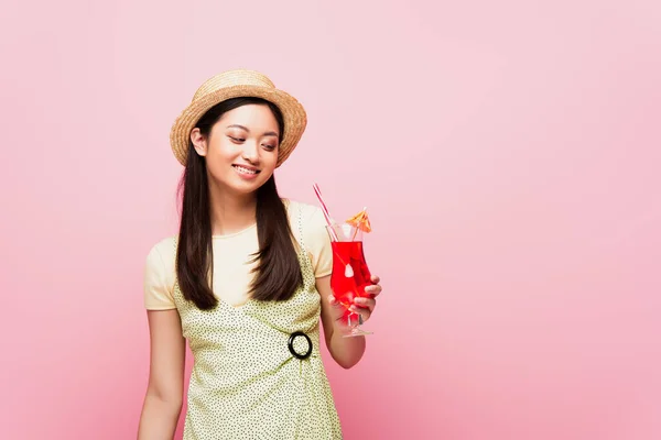 Smiling young asian woman in straw hat looking at glass with cocktail isolated on pink — Stock Photo