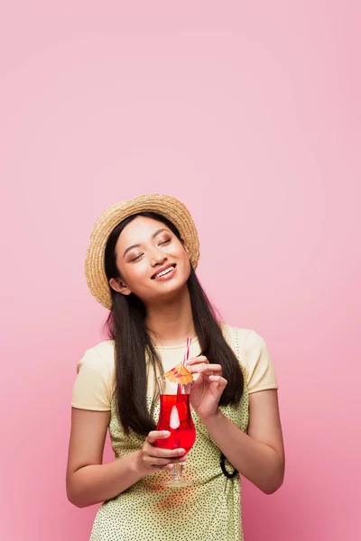 Sonriente joven asiática mujer con los ojos cerrados celebración de vidrio con cóctel aislado en rosa - foto de stock