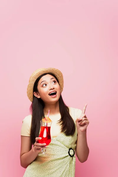Усміхнена молода азіатка в солом'яному капелюсі тримає склянку з коктейлем і вказує пальцем, дивлячись вгору ізольовано на рожевому — стокове фото