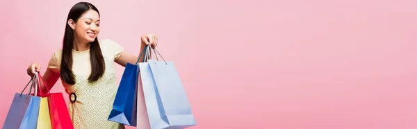 Concetto panoramico di giovane donna asiatica sorridente guardando borse della spesa isolate sul rosa — Foto stock