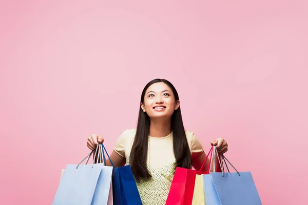 Alegre jovem asiático mulher olhando para cima enquanto segurando compras sacos isolado no rosa — Fotografia de Stock