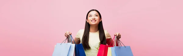 Horizontales Bild einer fröhlichen jungen asiatischen Frau, die aufblickt, während sie Einkaufstüten isoliert auf rosa hält — Stockfoto