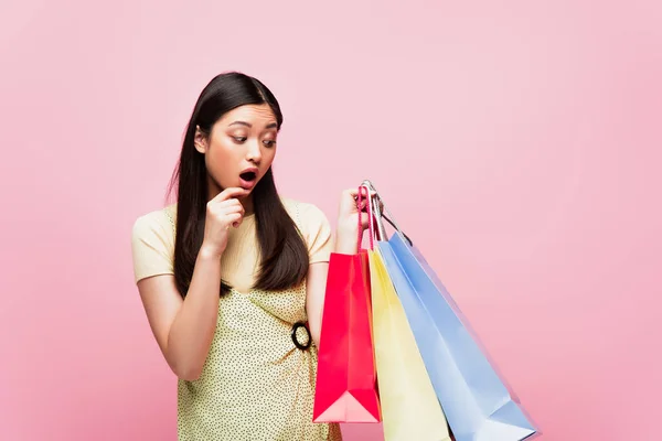 Schockiertes asiatisches Mädchen schaut auf Einkaufstüten isoliert auf rosa — Stockfoto