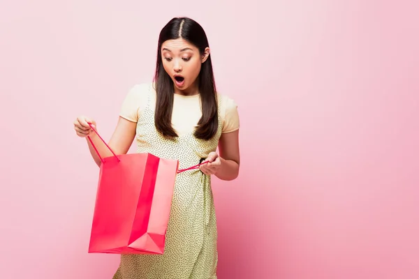 Impactado joven asiático mujer mirando bolsa de compras en rosa - foto de stock