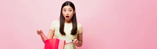 Panoramic crop of shocked young asian woman holding shopping bag on pink — Stock Photo