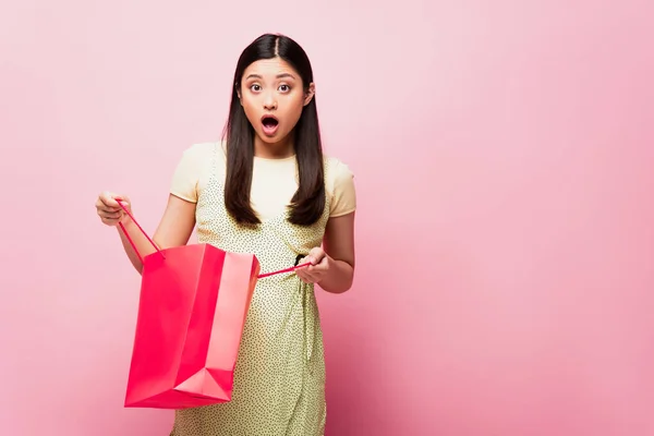 Impactado joven asiática mujer sosteniendo bolsa de compras en rosa - foto de stock